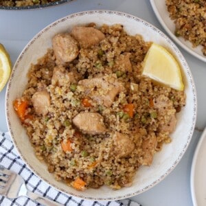 A bowl of quinoa mixed with chunks of cooked chicken, diced carrots, peas, and spices, garnished with a lemon wedge. The bowl is placed on a gray surface with a striped napkin beside it.