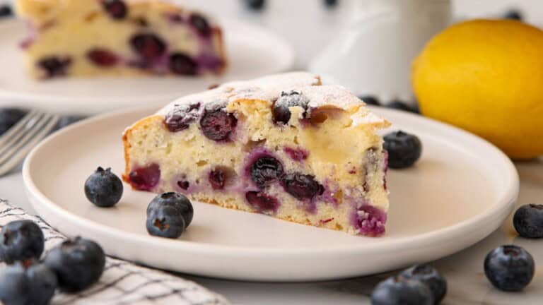 A slice of a simple blueberry cake on a white plate, surrounded by fresh blueberries and a lemon, with another plate of cake and a fork in the background. The cake is moist with visible blueberries and a light dusting of powdered sugar.