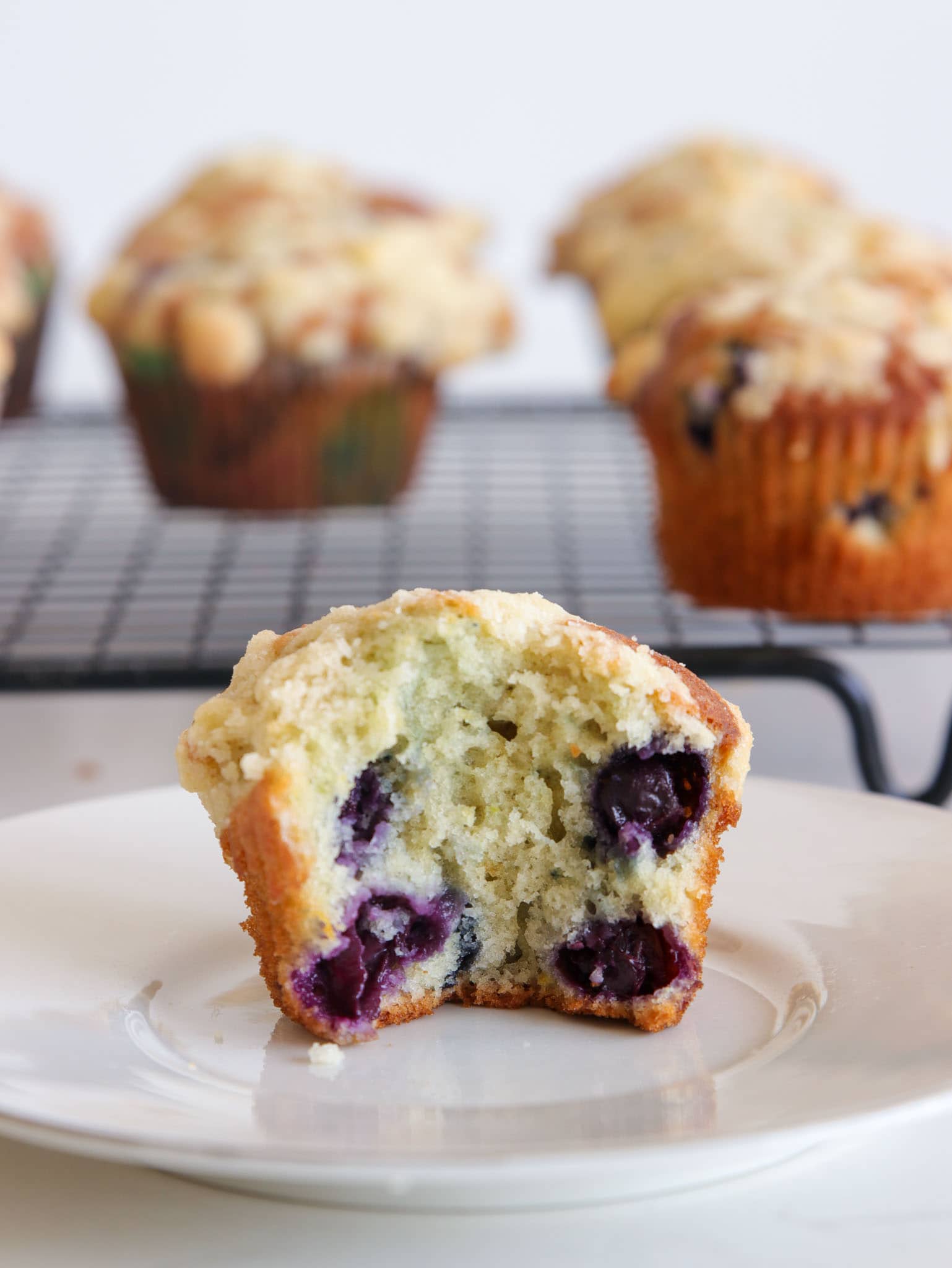 A blueberry muffin cut in half on a white plate, showing juicy blueberries inside. More muffins are cooling on a wire rack in the background.