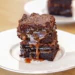 A close-up of a stack of rich chocolate brownies with gooey caramel sauce oozing between the layers, served on a white plate. Another plate with brownies is blurred in the background.