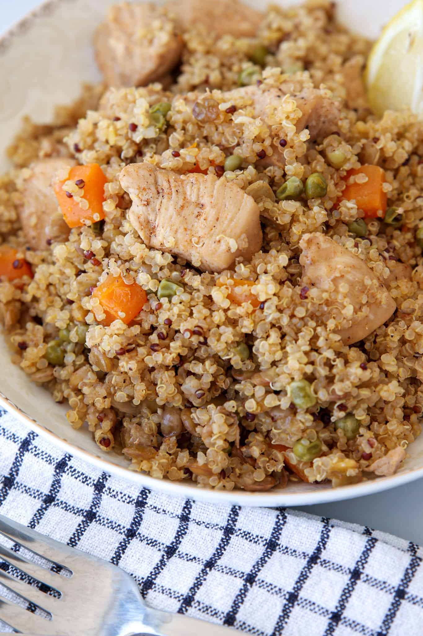 A close-up of a bowl filled with quinoa mixed with chunks of cooked chicken, diced carrots, peas, and seasoning, with a lemon wedge on the side. The bowl rests on a checkered cloth next to a fork.