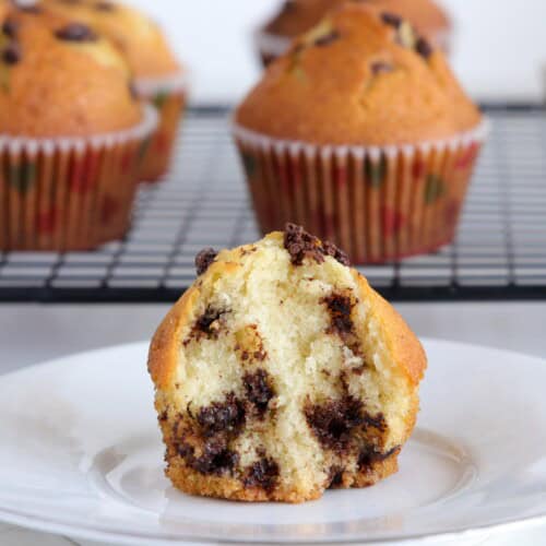 A chocolate chip muffin cut in half is on a white plate, with whole muffins in paper liners cooling on a black wire rack in the background.