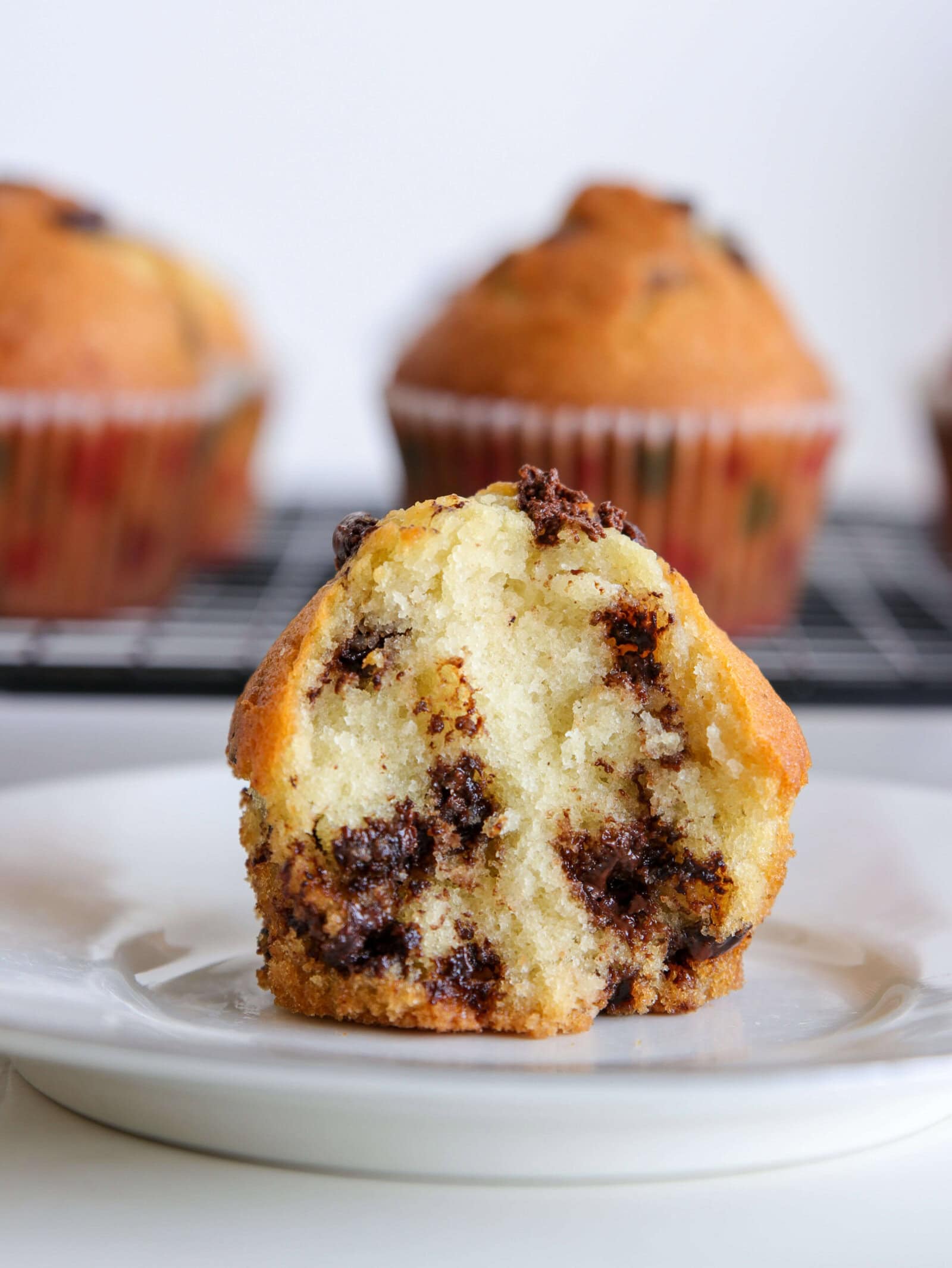 A chocolate chip muffin cut in half sits on a white plate, revealing a moist, fluffy inside with melted chocolate chips. Whole muffins on a cooling rack are blurred in the background.
