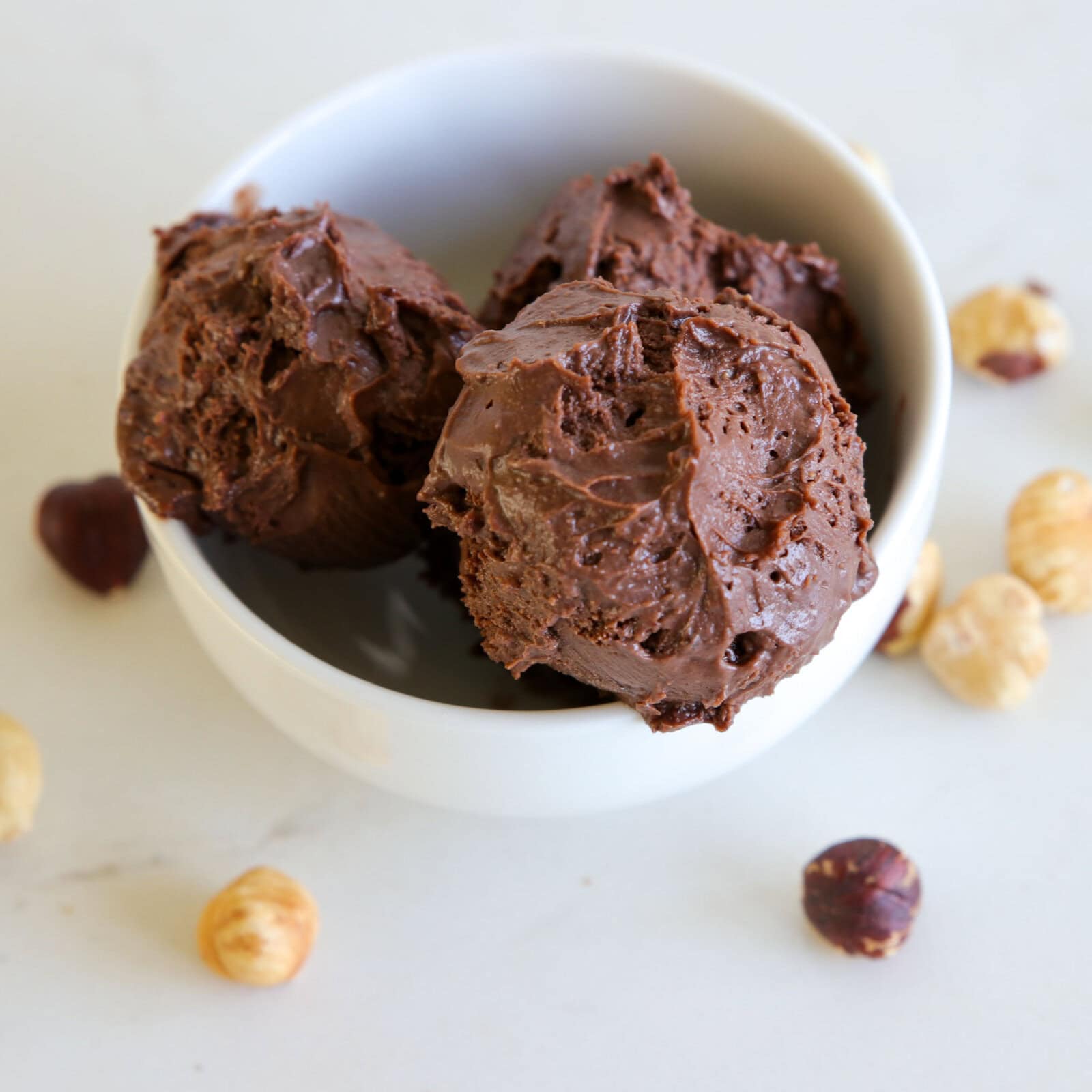 A white bowl filled with three scoops of Nutella ice cream sits on a white surface, surrounded by scattered hazelnuts.