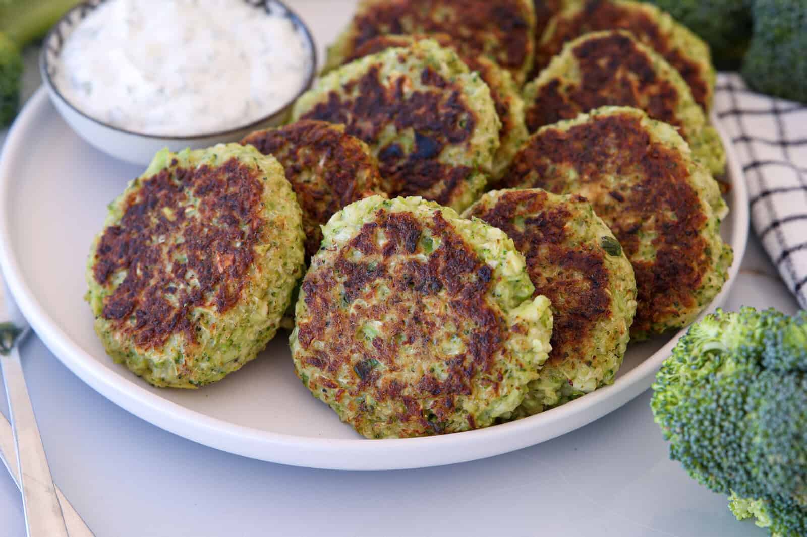 A plate filled with several golden-brown broccoli fritters, served alongside a small bowl of creamy dipping sauce. Fresh broccoli florets and a checkered napkin are visible nearby.