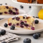 A slice of blueberry cake on a white plate, garnished with fresh blueberries and a dusting of powdered sugar; a whole lemon and another cake slice are in the background.