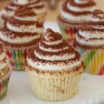 Several tiramisu cupcakes with swirled white frosting and a dusting of cocoa powder on top, displayed on a white plate. The cupcakes are in colorful, striped paper liners.