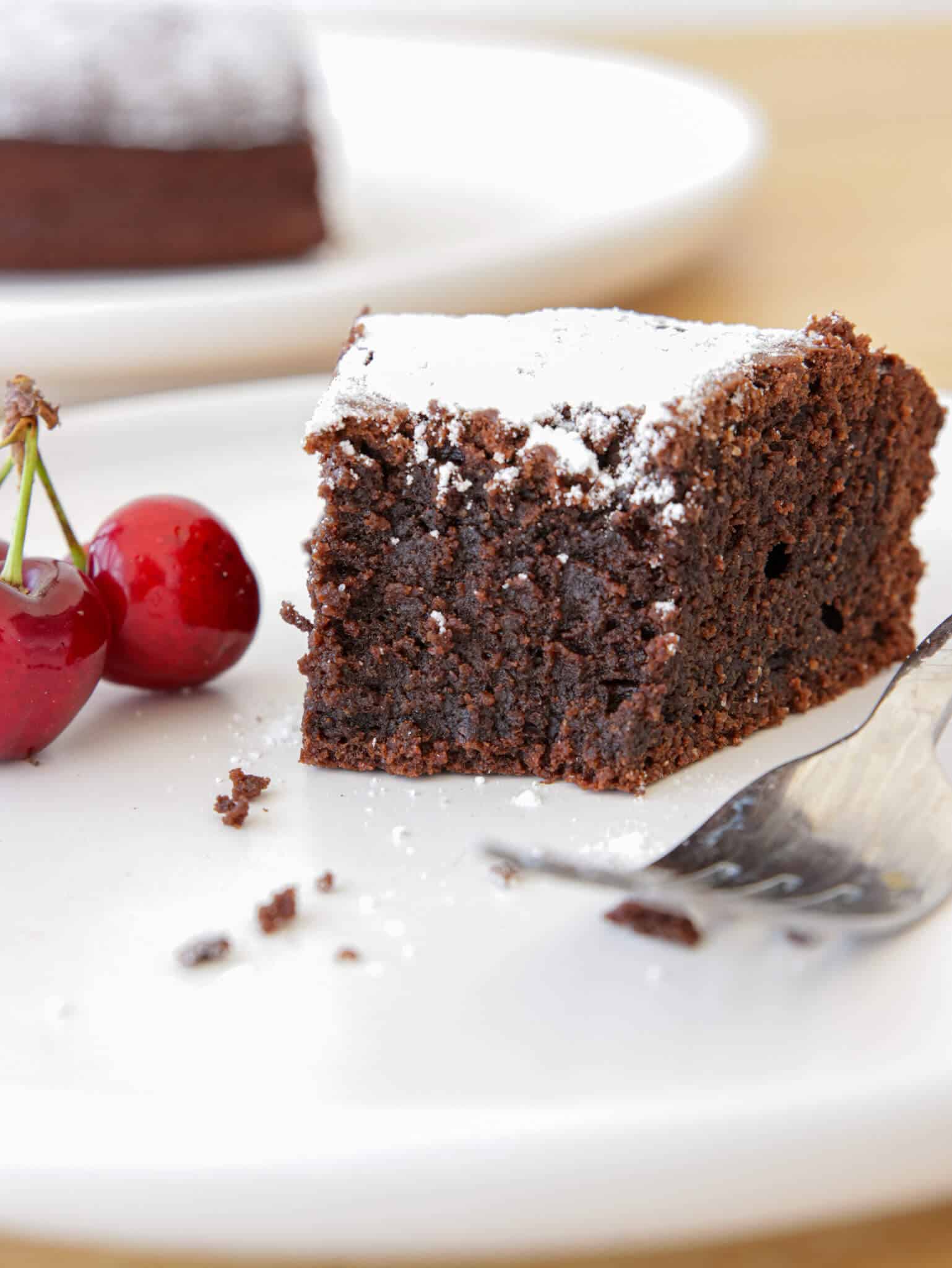 A slice of moist chocolate almond cake topped with powdered sugar on a white plate, next to two fresh cherries and a fork with cake crumbs around.