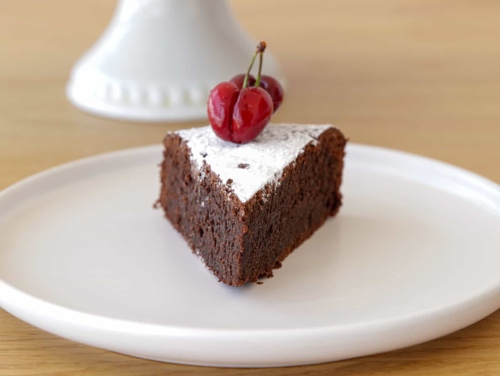 A slice of flourless chocolate almond cake topped with powdered sugar and two cherries sits on a white plate, with a cake stand blurred in the background.