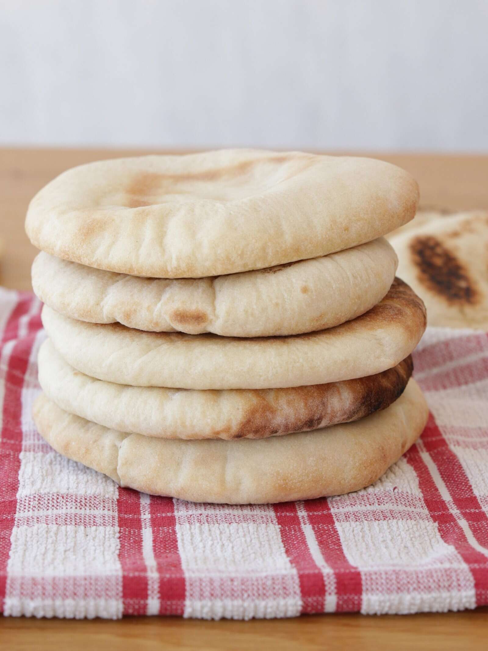A stack of round, fluffy pita breads sits on a red and white checkered kitchen towel on a wooden surface.