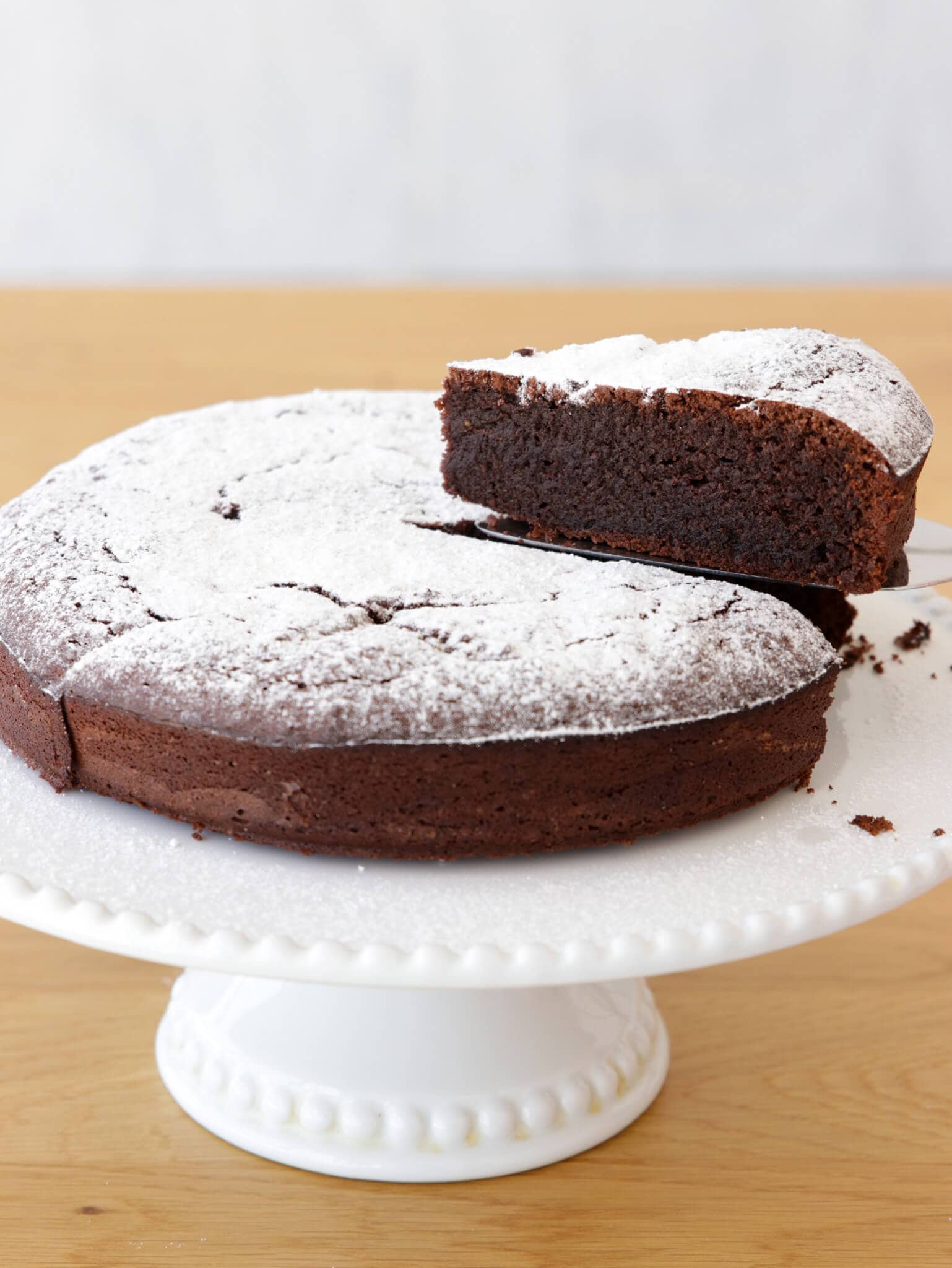 A round chocolate cake dusted with powdered sugar sits on a white cake stand. A single slice is being lifted, showing the cake's moist, rich texture inside.