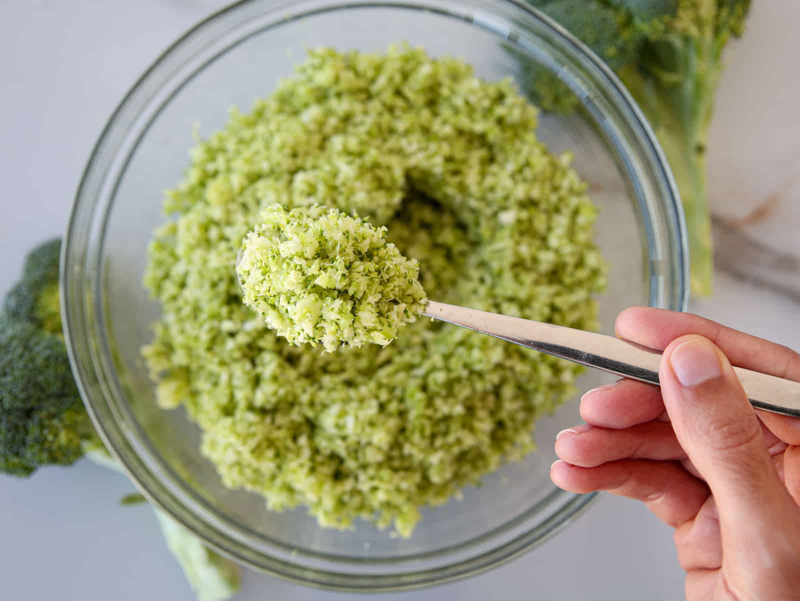 A hand holding a spoonful of finely chopped broccoli over a glass bowl filled with more chopped broccoli, with fresh broccoli in the background.