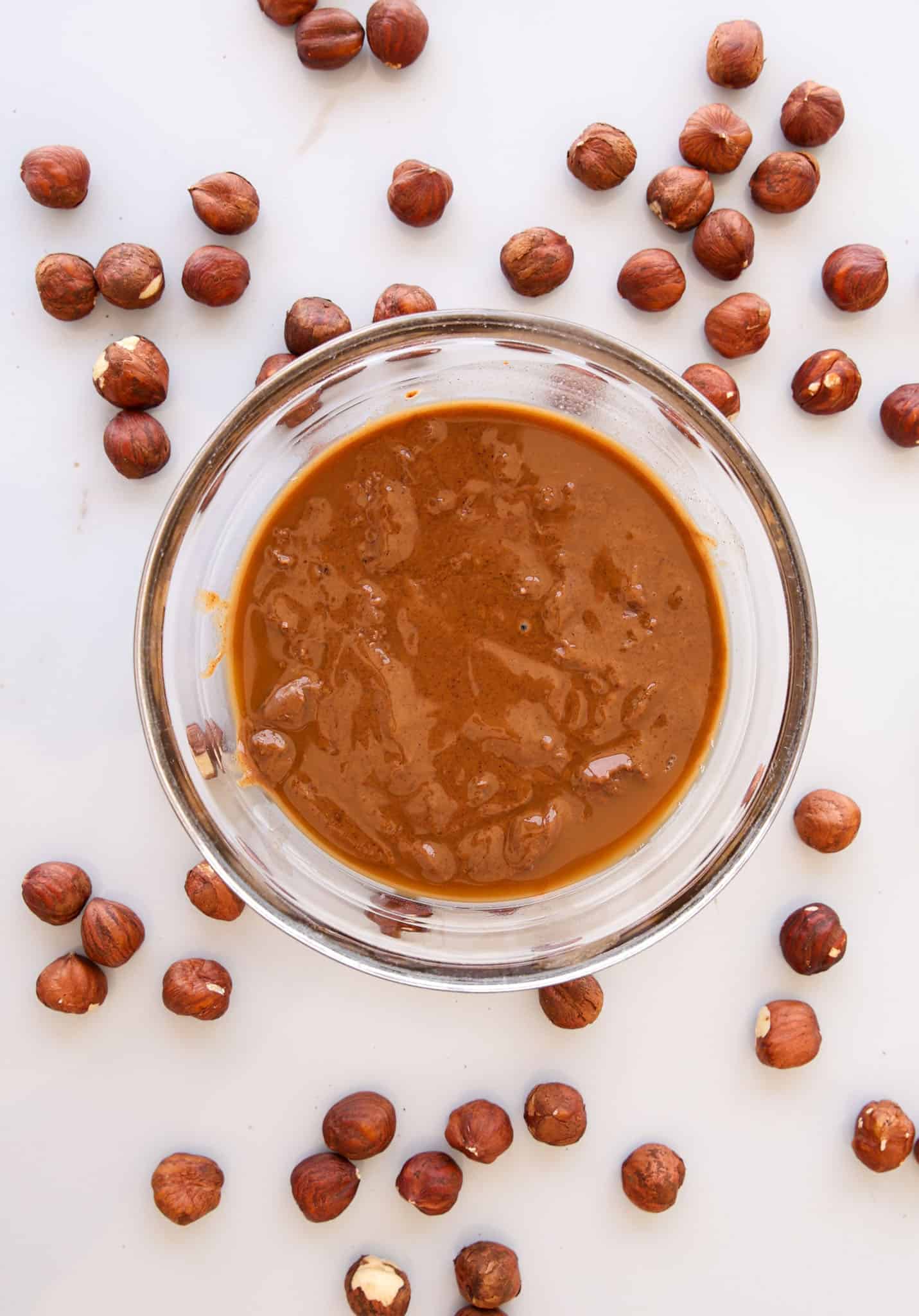 A glass bowl filled with creamy hazelnut spread is surrounded by scattered whole hazelnuts on a white surface.