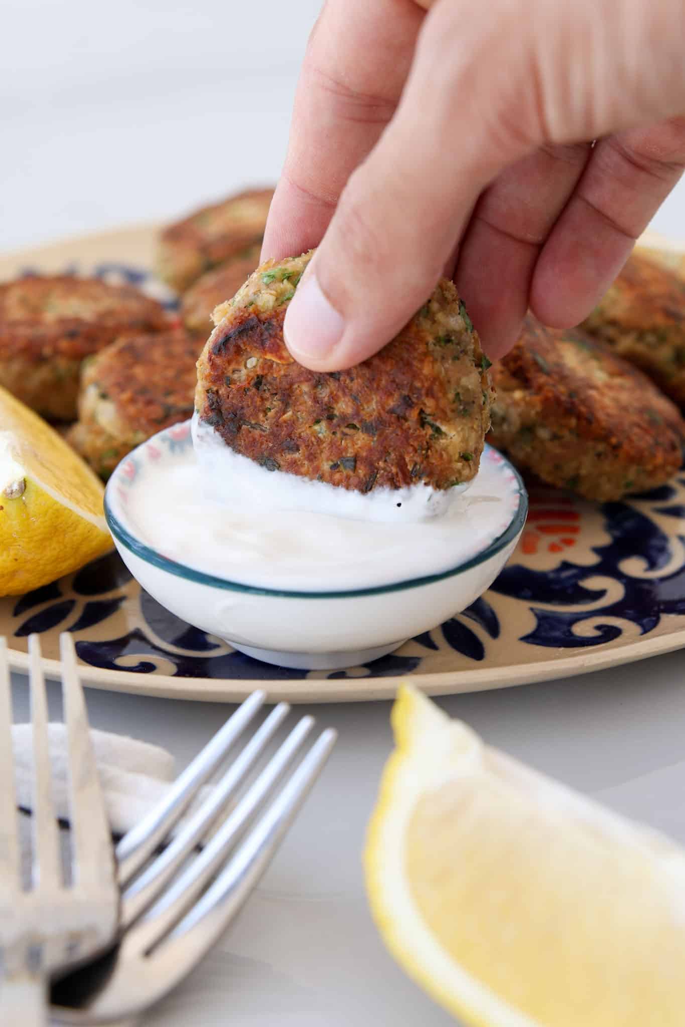 A hand dips a tuna oatmeal patty into a small bowl of white sauce, with more falafel and lemon wedges on a patterned plate in the background. Two forks and a napkin are in the foreground.