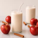 Two glasses of creamy apple pie smoothie, one with a glass straw, surrounded by red apples and cinnamon sticks on a white surface with a light background.