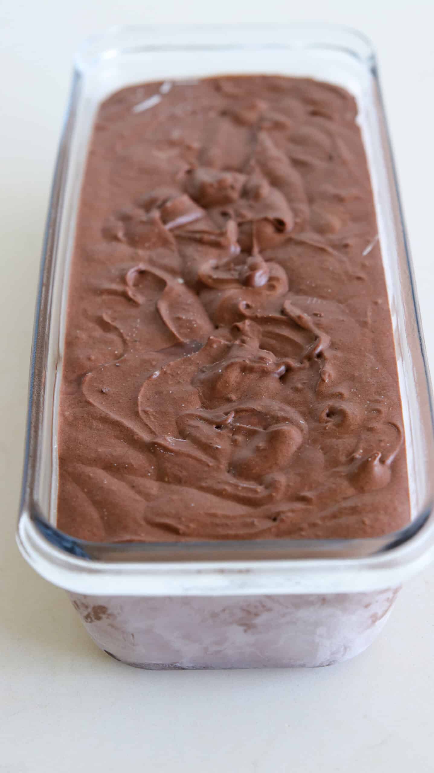 A glass container filled with smooth, swirled chocolate hazelnut ice cream sits on a light surface, viewed from above and slightly to the side.