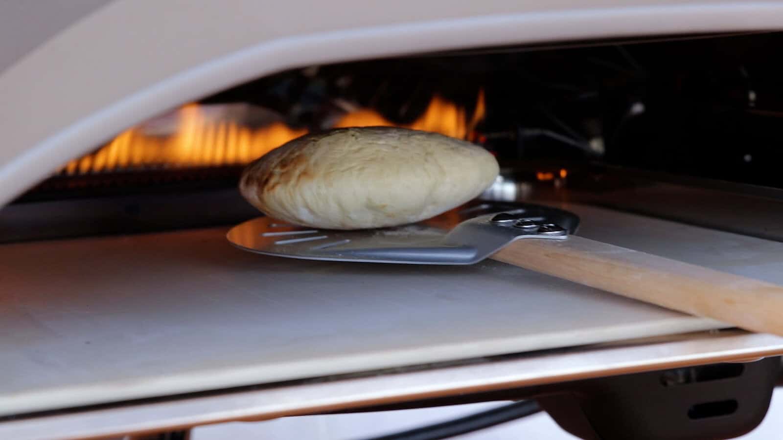 A round, puffed pita bread is being removed from a hot oven using a metal pizza peel with a wooden handle. Flames are visible in the background inside the oven.