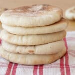 A stack of six round pita breads sits on a red and white checkered cloth, with two more pita breads blurred in the background.