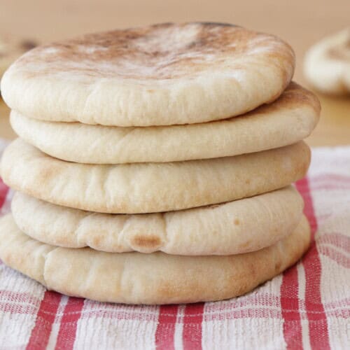 A stack of six round pita breads sits on a red and white checkered cloth, with two more pita breads blurred in the background.