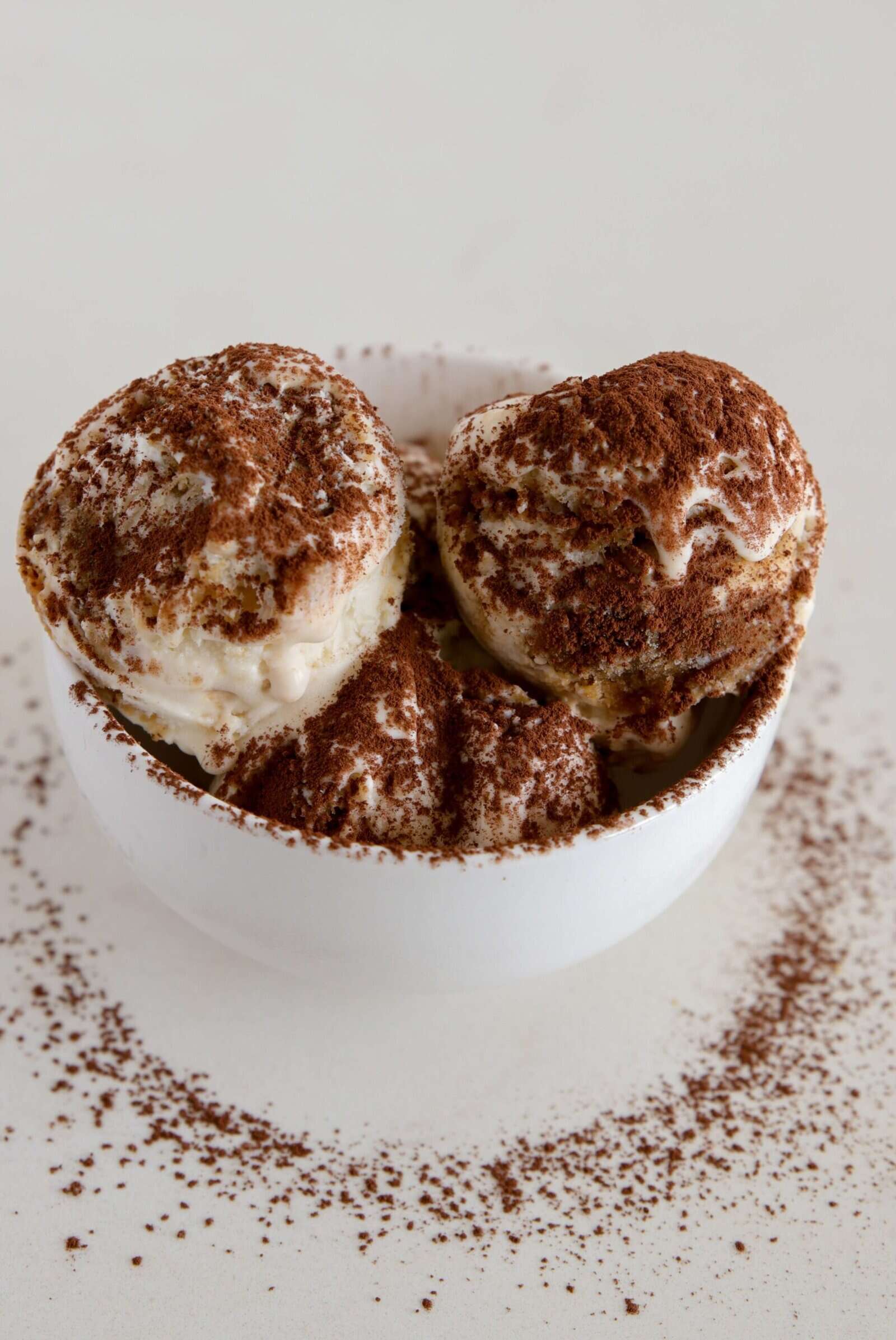 A white bowl filled with three scoops of vanilla ice cream is dusted with cocoa powder, with extra cocoa sprinkled on the surface around the bowl.