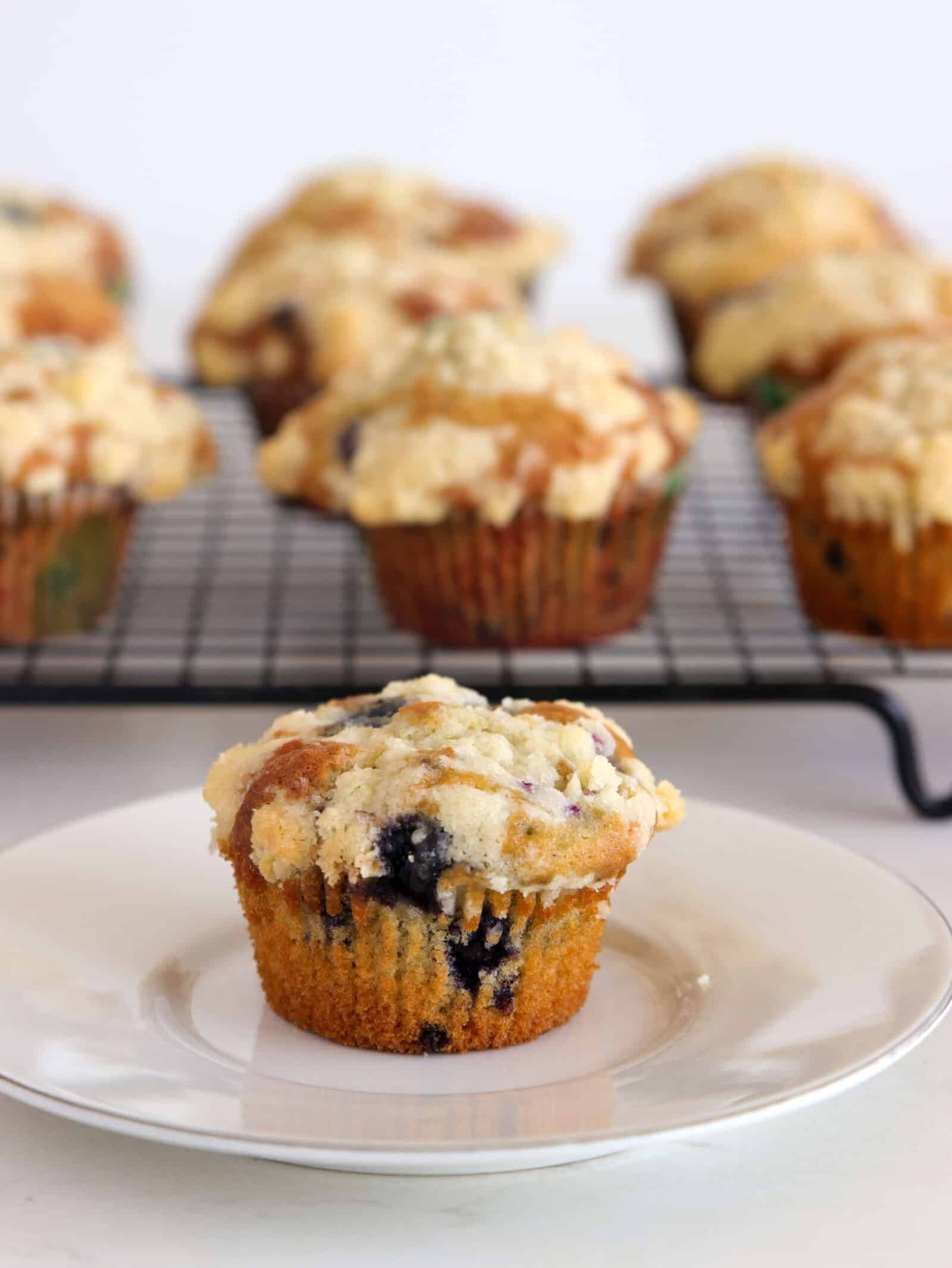 A blueberry muffin with crumb topping sits on a white plate, with more muffins cooling on a wire rack in the background.