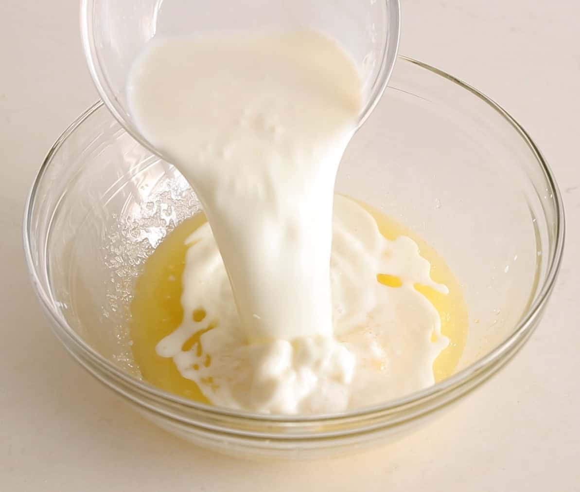 A clear glass bowl with a yellow liquid inside, while a stream of buttermilk is being poured into it from a smaller glass container above.