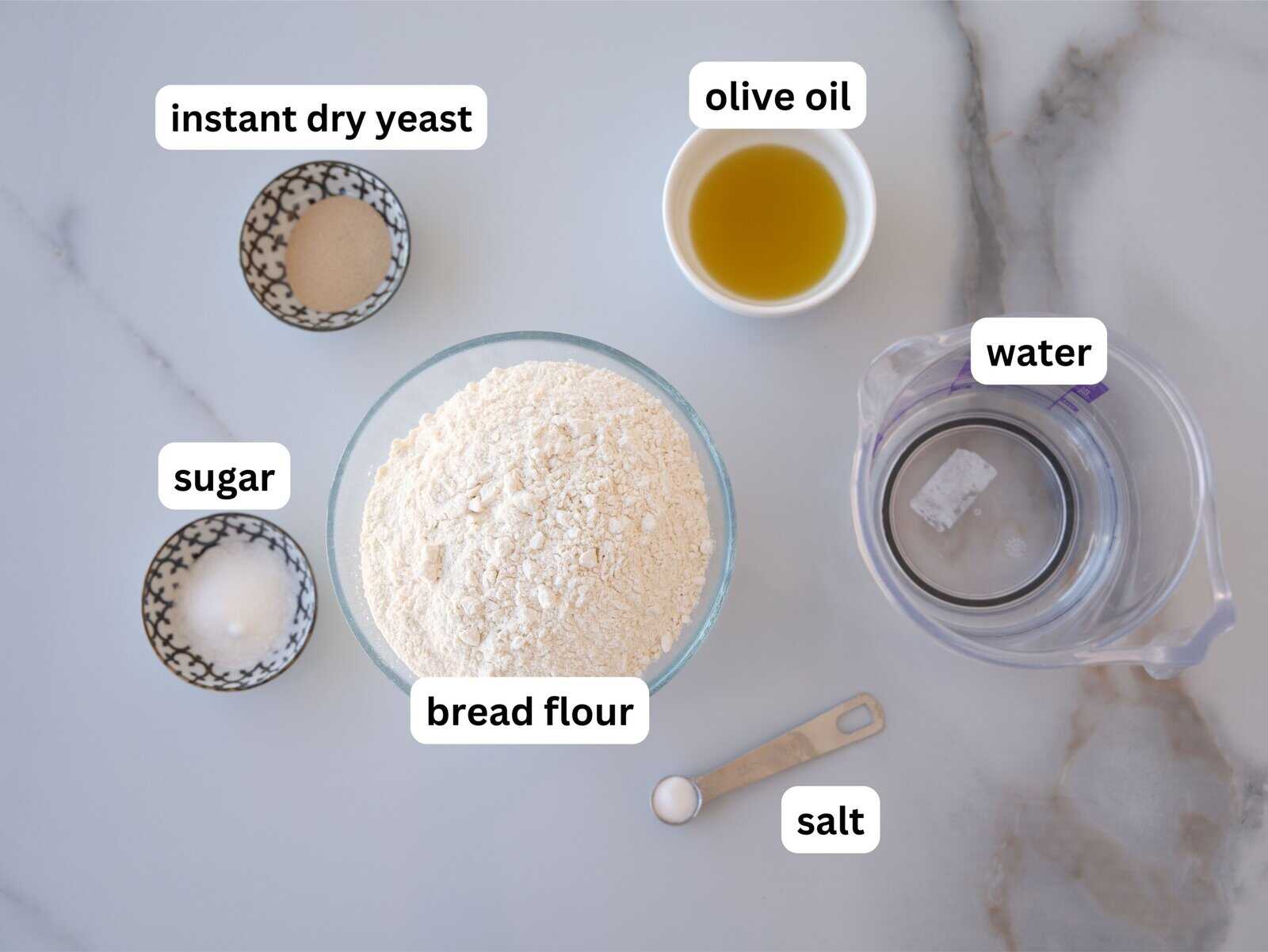 Top view of labeled ingredients for bread: instant dry yeast, bread flour, sugar, salt, olive oil, and a jug of water on a white marble surface.