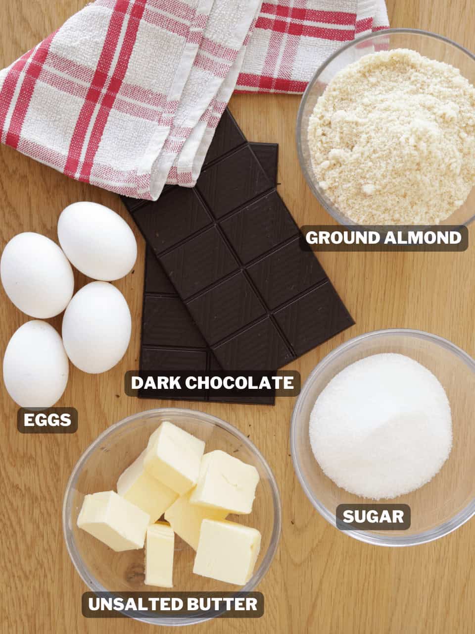 A top-down view of baking ingredients: a red and white towel, four eggs, a bar of dark chocolate, a bowl of ground almond, a bowl of sugar, and a bowl of unsalted butter, all labeled.