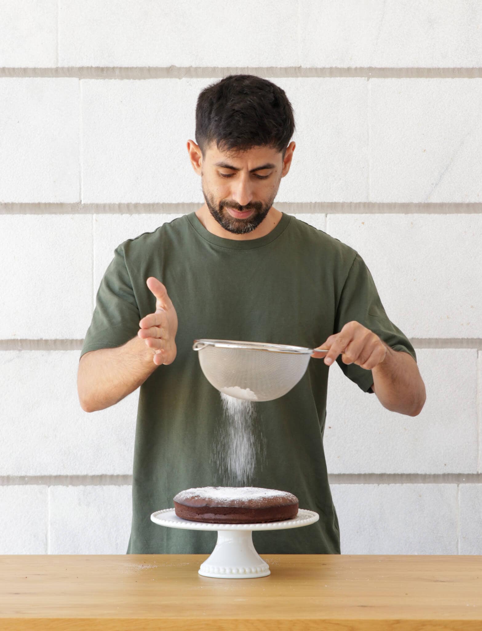 David in a green t-shirt sprinkles powdered sugar onto a chocolate cake using a sieve, standing behind a wooden table with a white brick wall in the background.
