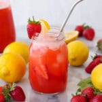 A mason jar filled with strawberry lemonade, ice, and fruit slices, garnished with a strawberry and lemon wedge on the rim. Whole strawberries and lemons are scattered on a white surface around the jar and a pitcher.