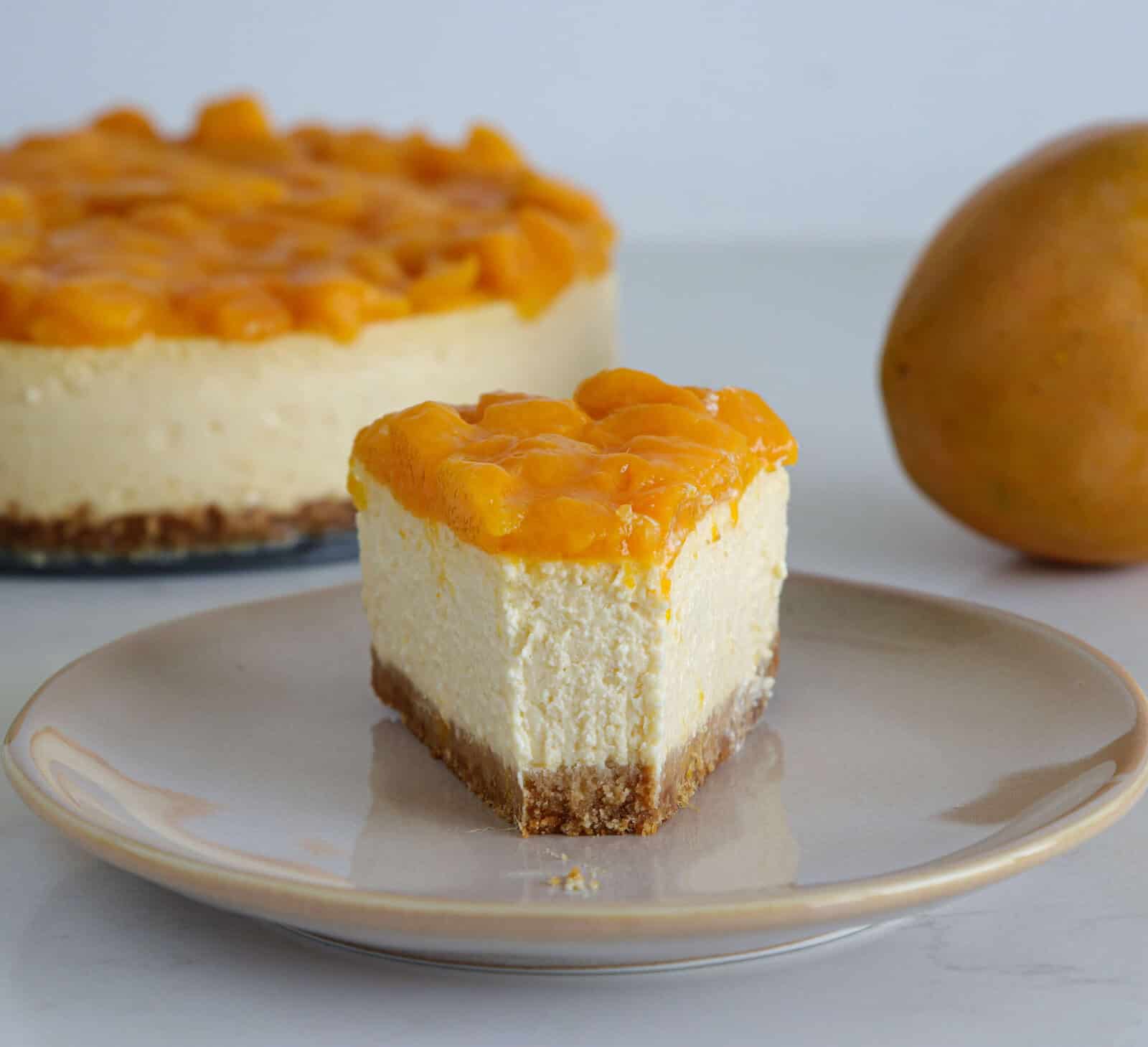 A slice of cheesecake topped with mango pieces sits on a plate, with the rest of the cheesecake and a whole mango in the background on a white surface.
