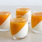 Four clear glasses filled with a dessert featuring a diagonal split of white panna cotta and bright mango puree, set on a light surface, The contrasting layers create a visually appealing pattern.
