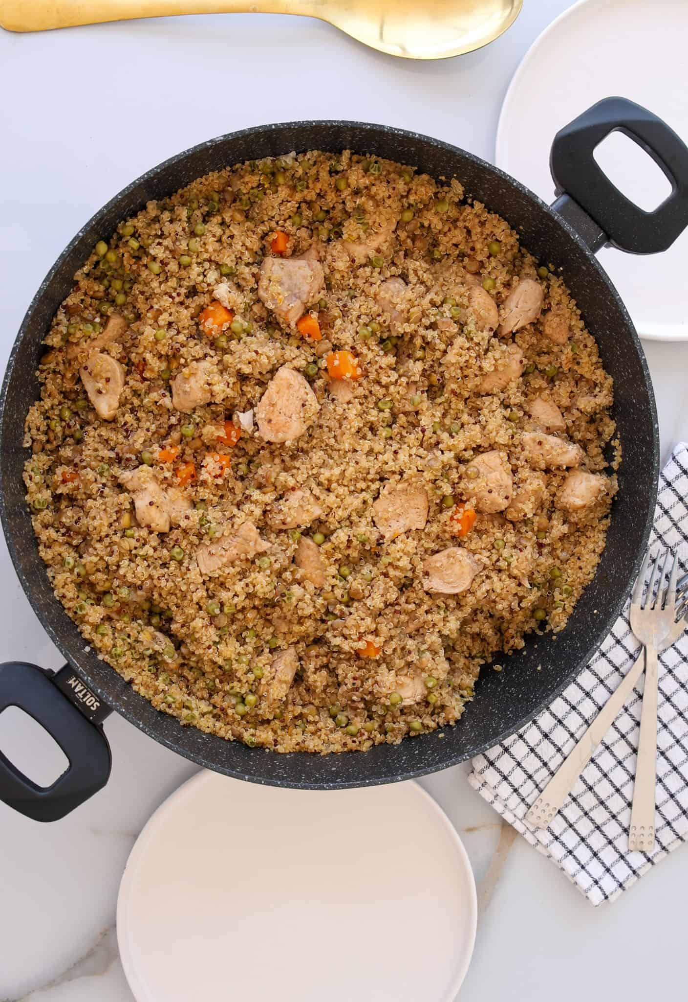 A large black skillet filled with quinoa, diced chicken, peas, and carrots sits on a white surface next to an empty plate, a fork, a striped napkin, and a serving spoon.