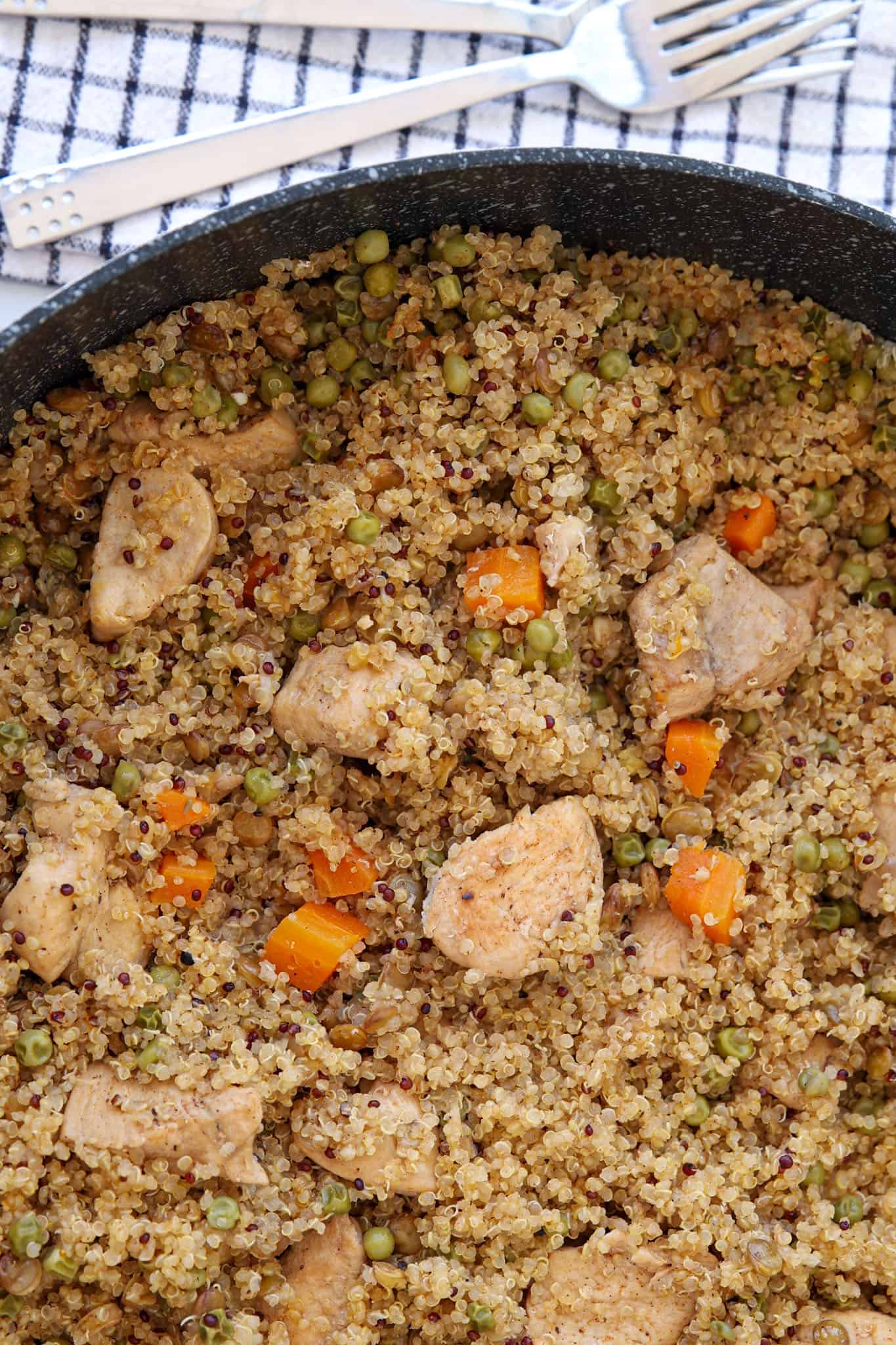 A skillet filled with quinoa, cooked chicken pieces, diced carrots, and green peas sits on a striped kitchen towel with a fork and knife beside it.