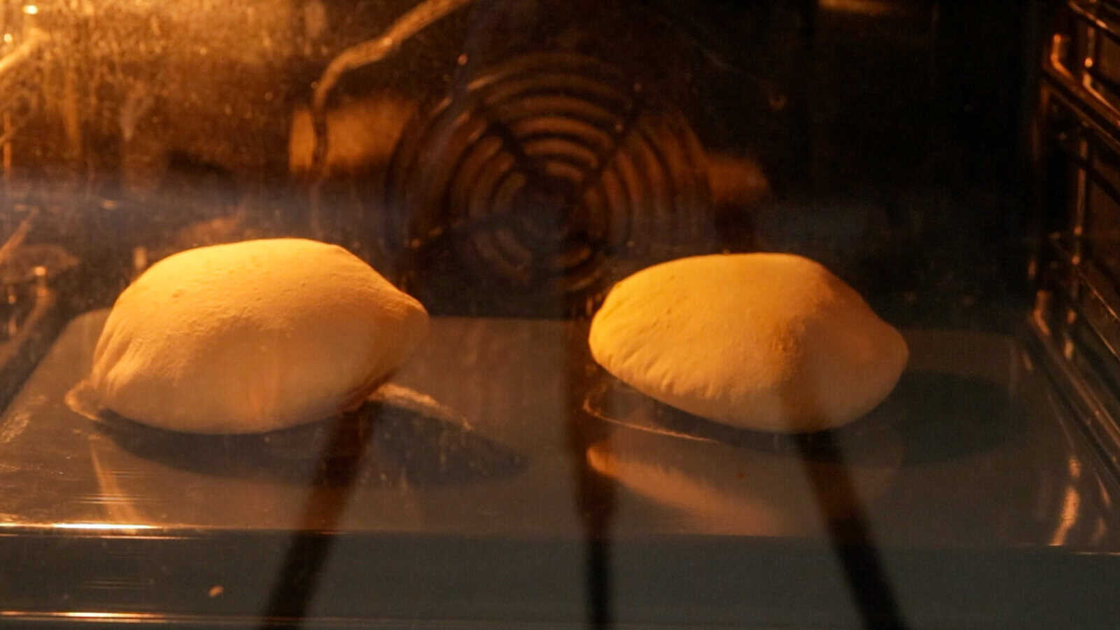 Two pieces of pita bread dough are puffing up and baking inside an oven, viewed through the oven door with an orange glow reflecting off the glass.
