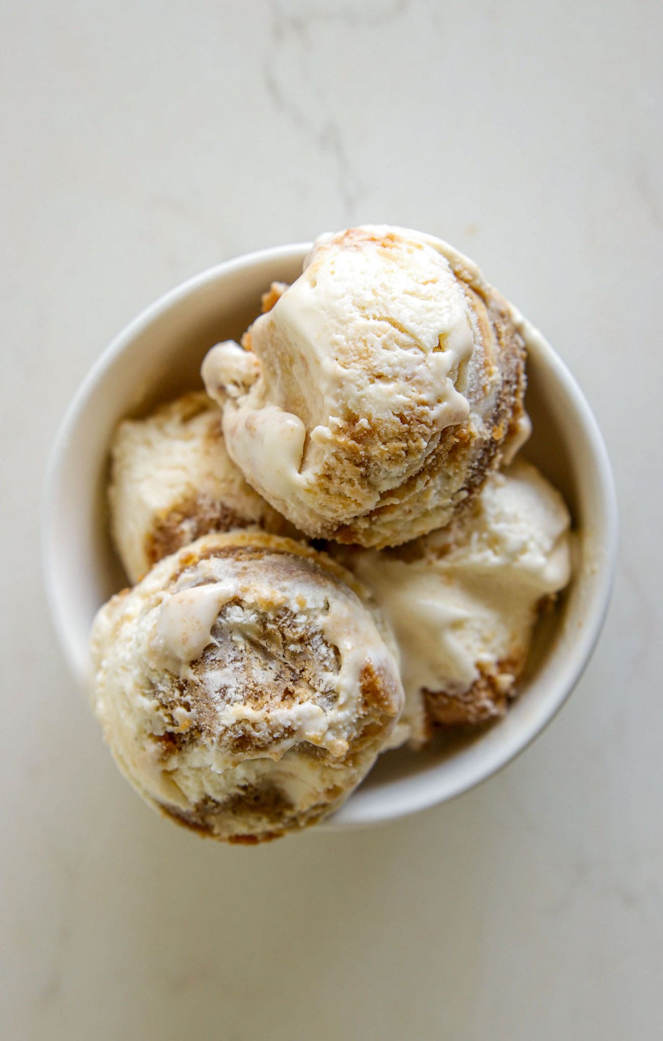A white bowl filled with three scoops of creamy ice cream with brown swirls, placed on a light-colored surface.