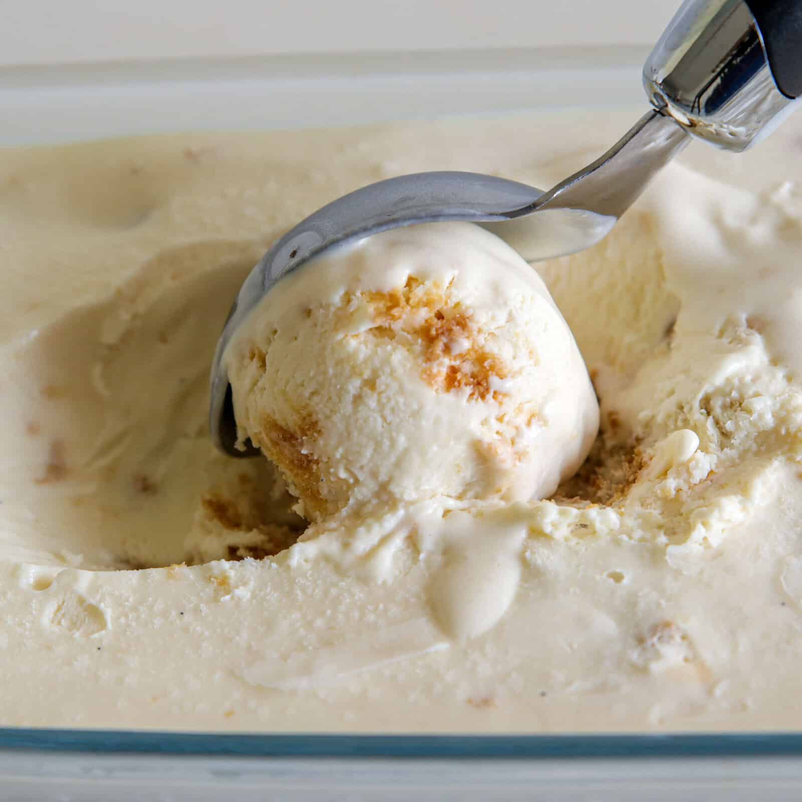 A metal ice cream scoop is lifting a round scoop of creamy, pale-colored ice cream with visible chunks from a rectangular container.
