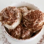 A white bowl filled with creamy scoops of tiramisu ice cream, generously dusted with cocoa powder, sits on a white surface with some cocoa sprinkled around.