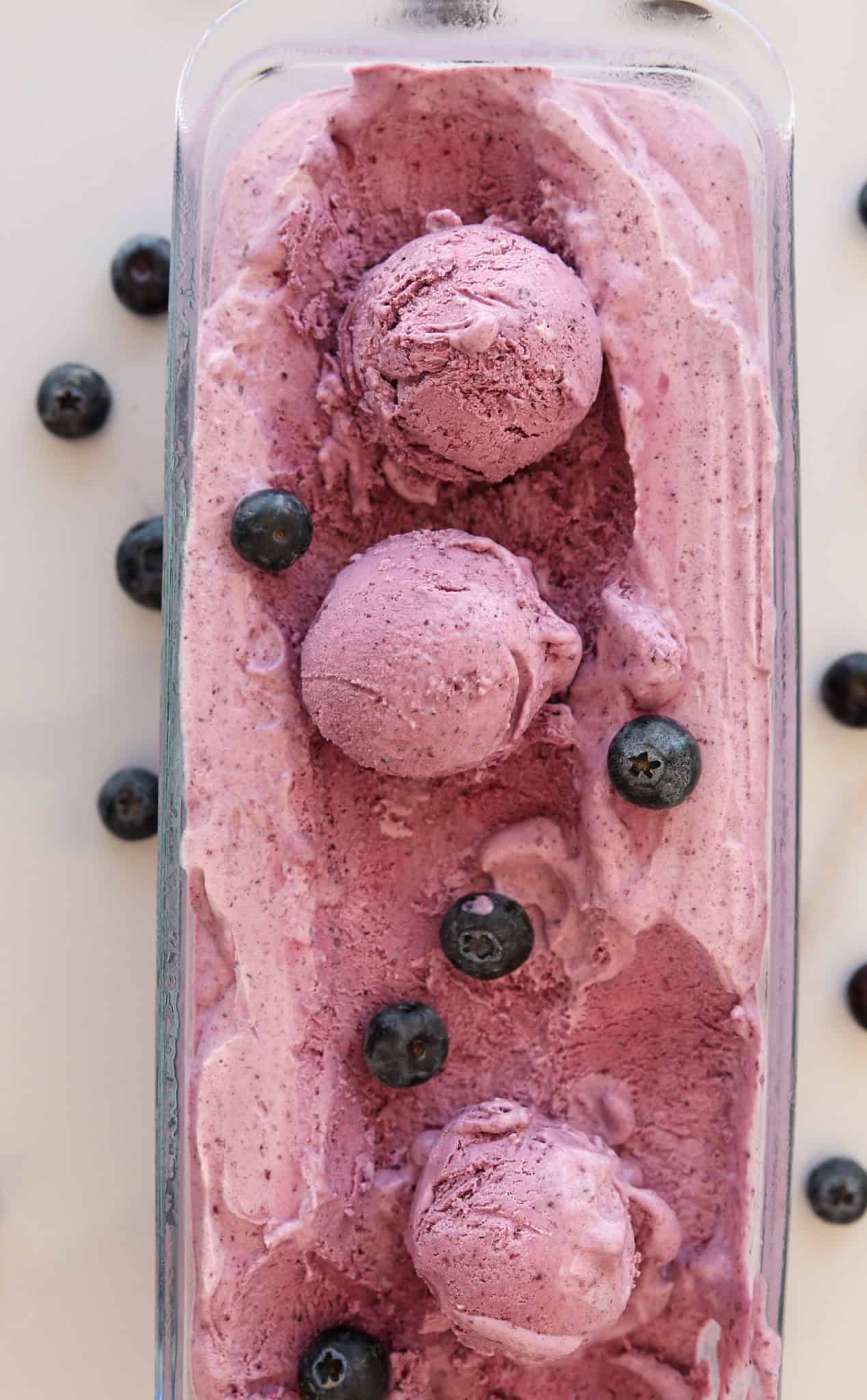 Rectangular glass dish filled with creamy purple blueberry ice cream, topped with three scoops and scattered fresh blueberries on top and around the dish.