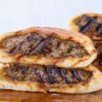 Two halves of grilled pita bread are stuffed with spiced ground meat and stacked on a wooden surface, showing the juicy, grilled filling inside; Another similar sandwich is visible in the background.