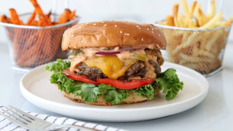 Best Burger Recipe With the Best Beef Blend Tips A cheeseburger with lettuce, tomato, onion, pickles, and special sauce on a sesame seed bun, served on a white plate with baskets of fries and sweet potato fries in the background.