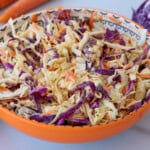 A colorful bowl of coleslaw with shredded green and purple cabbage and carrots, mixed with a creamy dressing. The bowl is orange and patterned, and fresh vegetables are visible in the background.