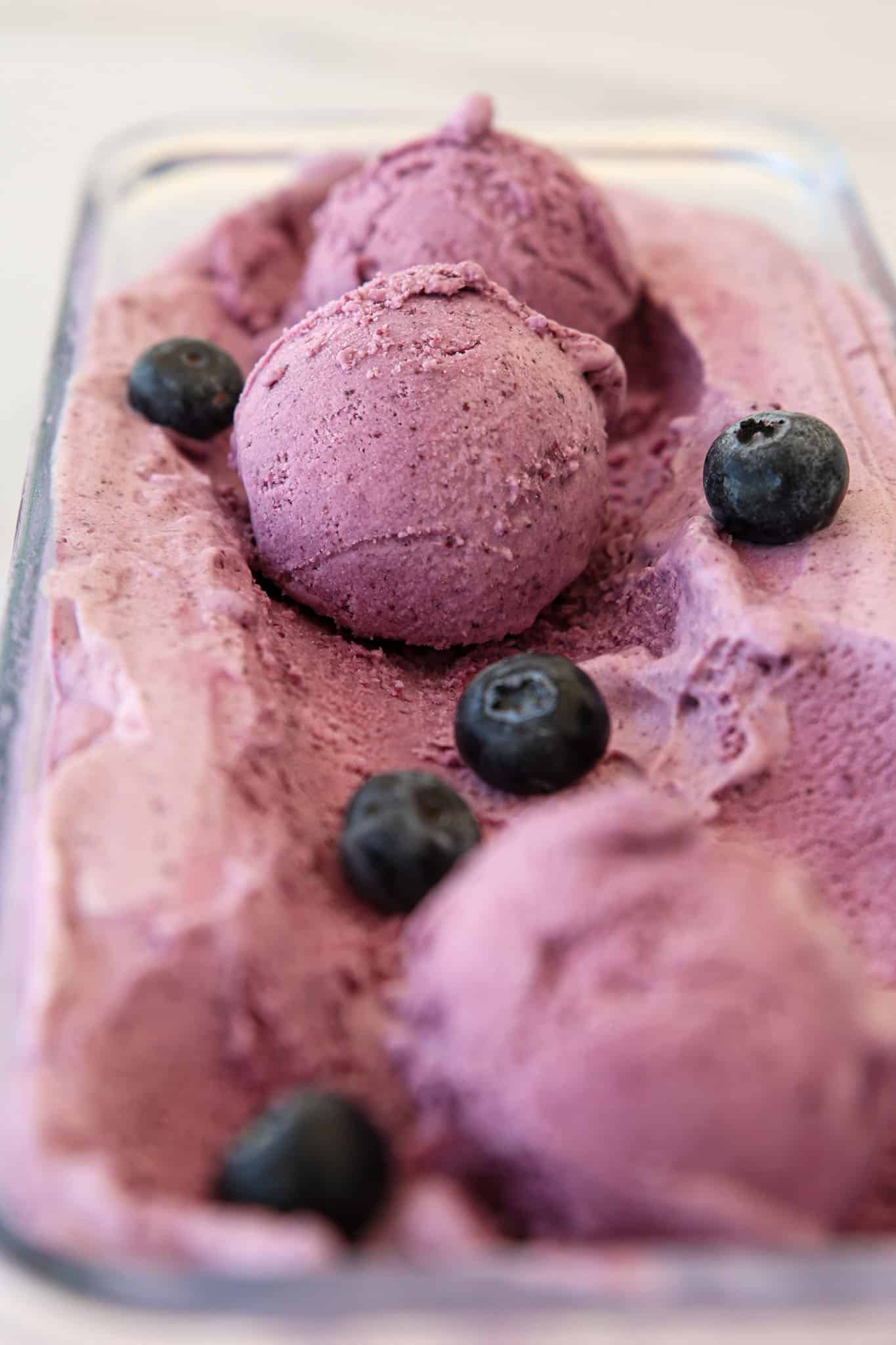 Close-up of a container with scoops of creamy blueberry ice cream, garnished with fresh whole blueberries.