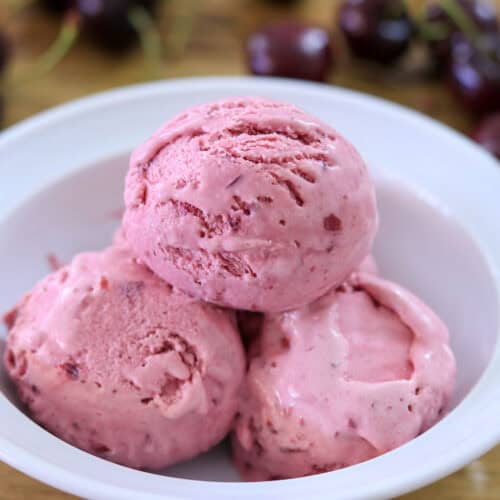 Three scoops of pink cherry ice cream in a white bowl, with fresh cherries blurred in the background on a wooden surface.