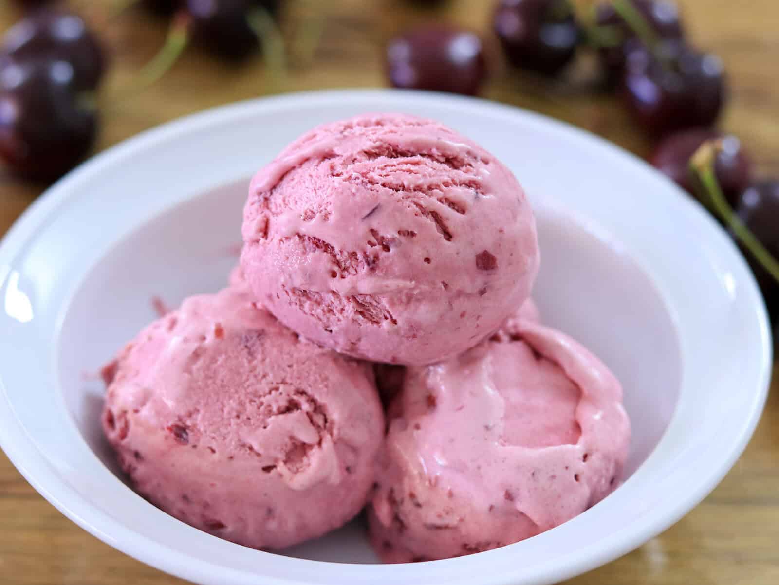 Three scoops of pink cherry ice cream in a white bowl, with fresh cherries blurred in the background on a wooden surface.