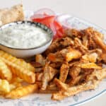 A plate with seasoned sliced chicken, crinkle-cut fries, pita bread, a bowl of creamy sauce, and fresh tomato and onion slices.