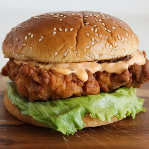 A close-up of a fried chicken sandwich with a sesame seed bun, crispy chicken fillet, fresh green leaf lettuce, and creamy sauce, placed on a wooden surface.