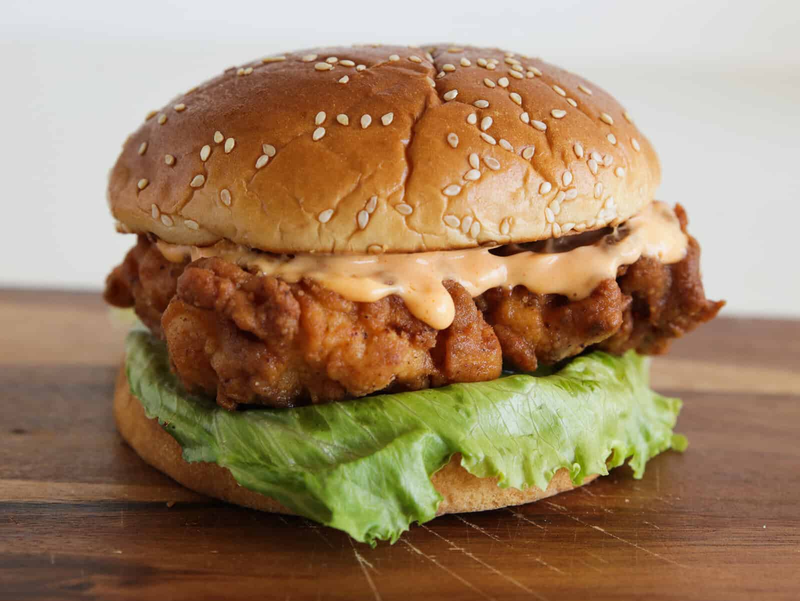 A close-up of a fried chicken sandwich with a sesame seed bun, crispy chicken fillet, fresh green leaf lettuce, and creamy sauce, placed on a wooden surface.