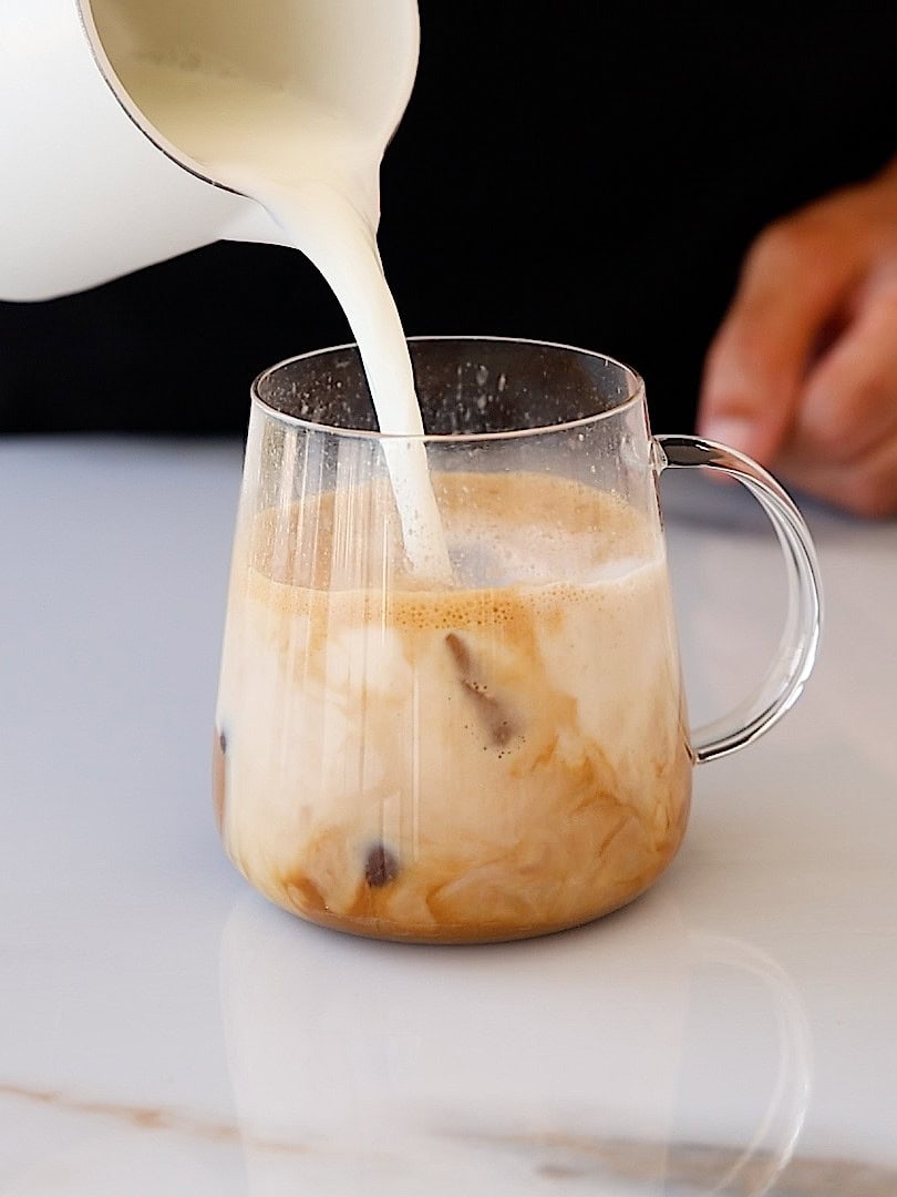 A hand pours creamy milk from a white pitcher into a glass of iced coffee on a white surface, creating swirling patterns in the drink.