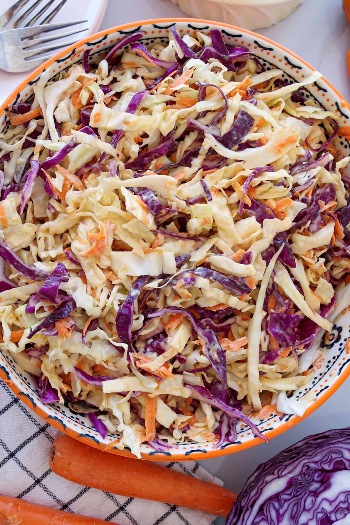 A close-up of a bowl filled with creamy coleslaw made of shredded green and purple cabbage and carrots, set on a patterned cloth with a fork, carrots, and a head of red cabbage nearby.